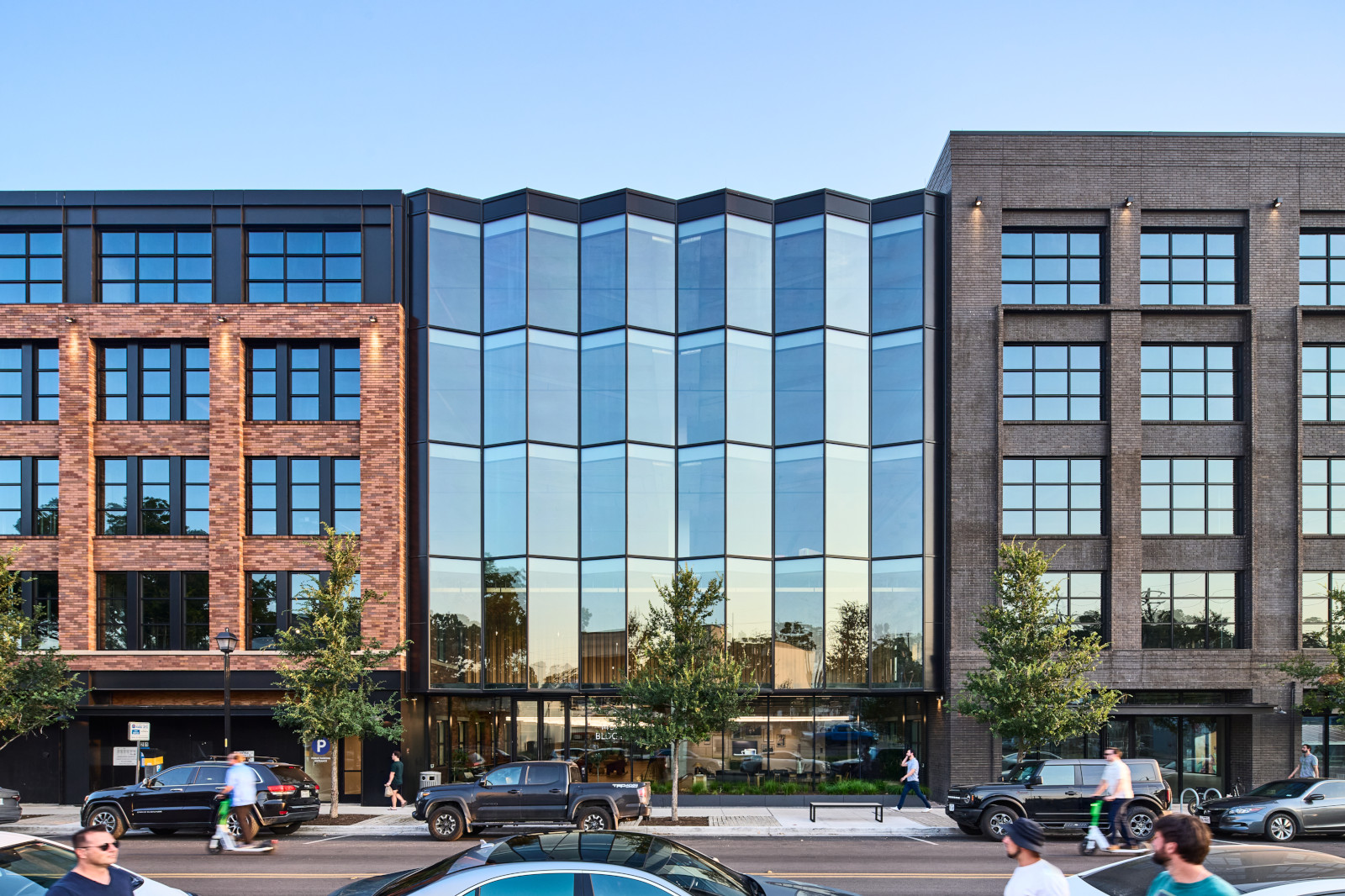 A modern building featuring a glass facade, flanked by brick structures, with pedestrians and vehicles in a vibrant urban setting.
