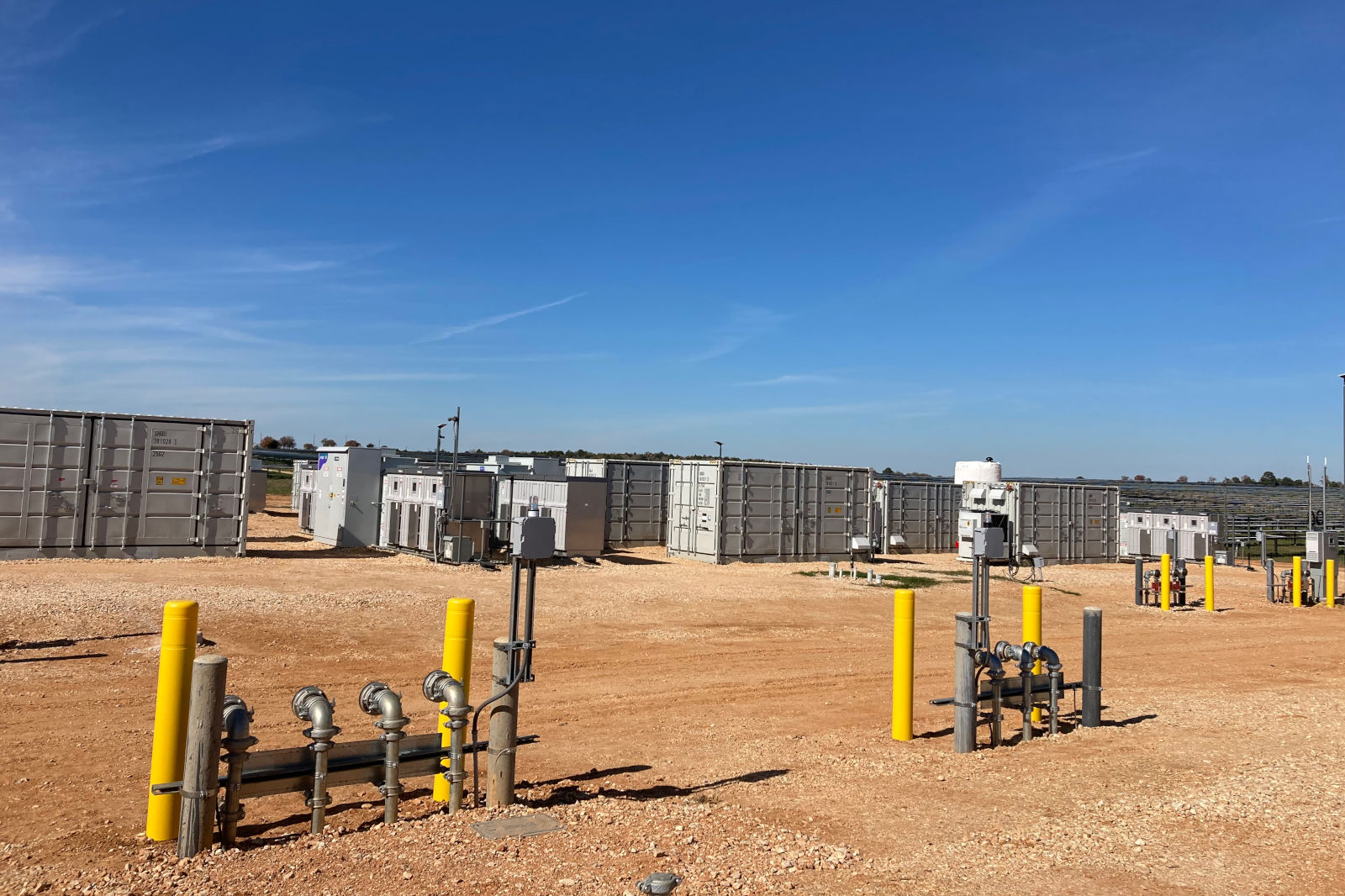 An industrial site with shipping containers, yellow barriers, and piping against a clear blue sky.