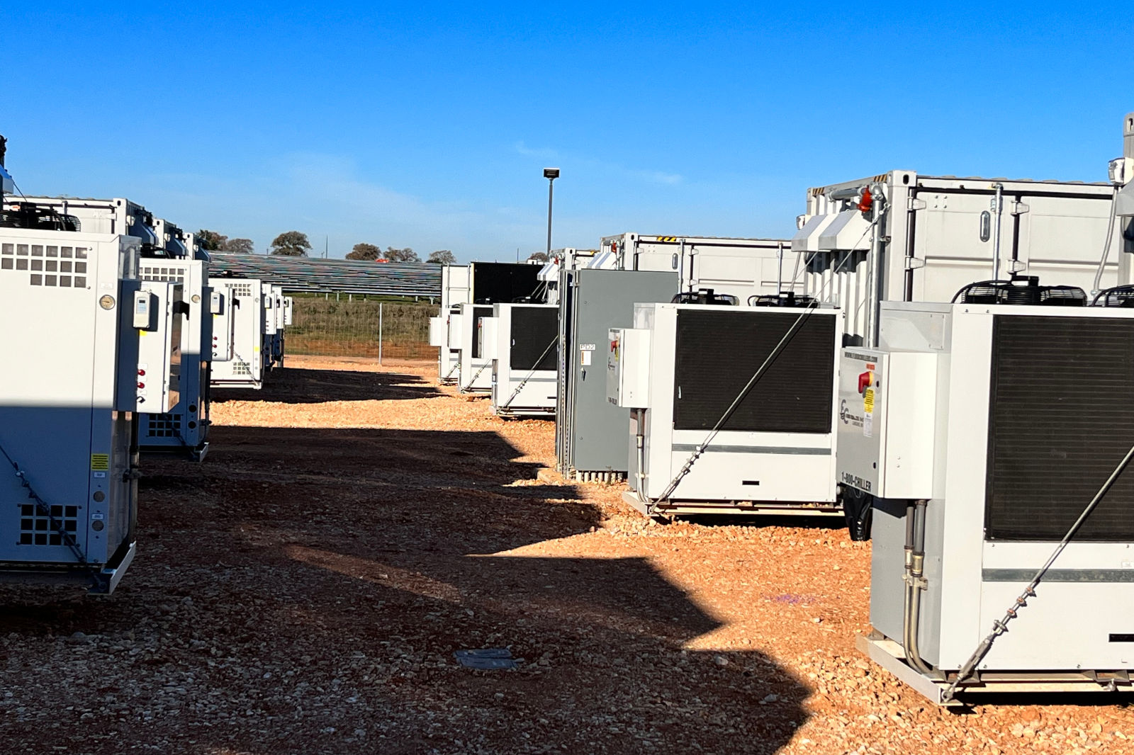 A row of large cooling units is set up on gravel ground, surrounded by a clear blue sky and distant trees.