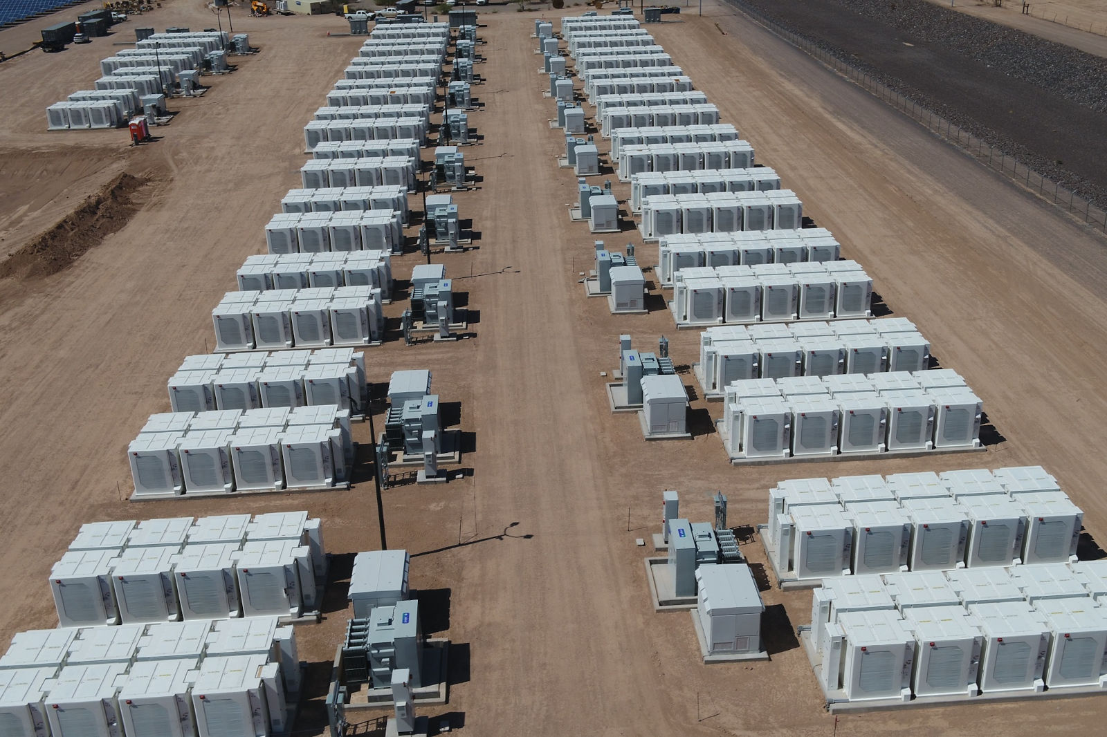 Aerial view of a large battery storage facility featuring rows of white energy storage units on a sandy terrain.