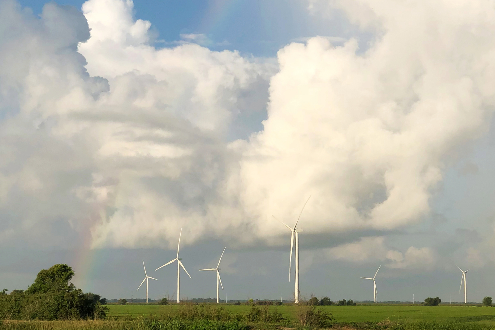 A landscape featuring wind turbines under a cloudy sky with a hint of a rainbow on the left side.