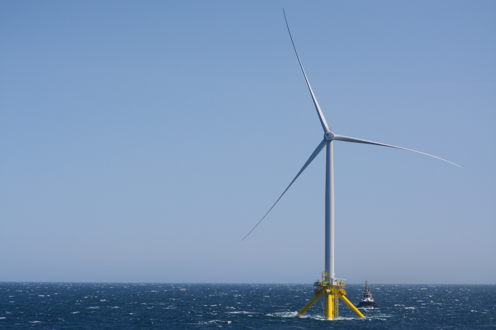 A large offshore wind turbine stands on a yellow platform in the ocean, with a small tugboat nearby.