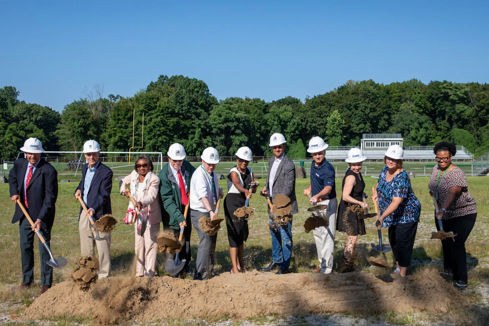 Group of individuals in hard hats holding shovels, participating in a groundbreaking ceremony on a sunny day.