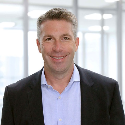 A professionally dressed man in a dark blazer and light blue shirt stands in an office setting, with blurred background.