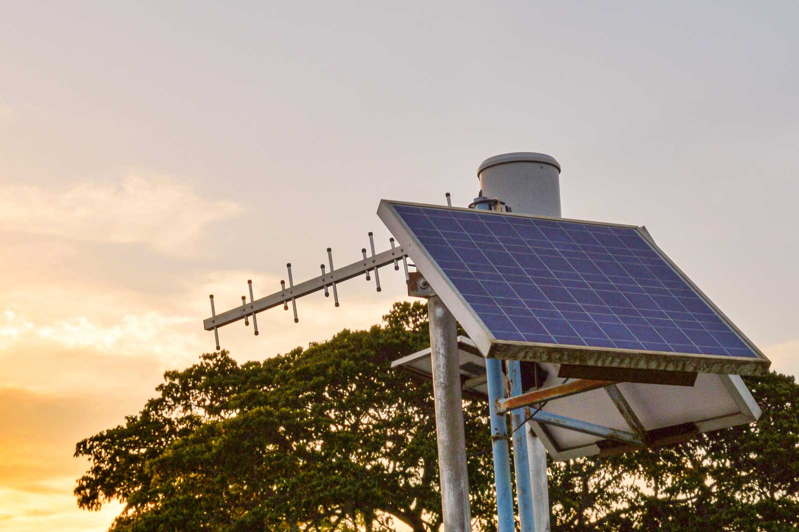 A solar panel mounted on a metal structure with antennas, set against a sunset background and lush trees.