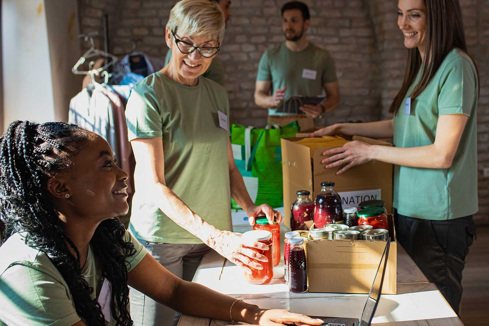 A group of volunteers in green shirts is organising food donations, with jars and boxes on a table in a well-lit room.