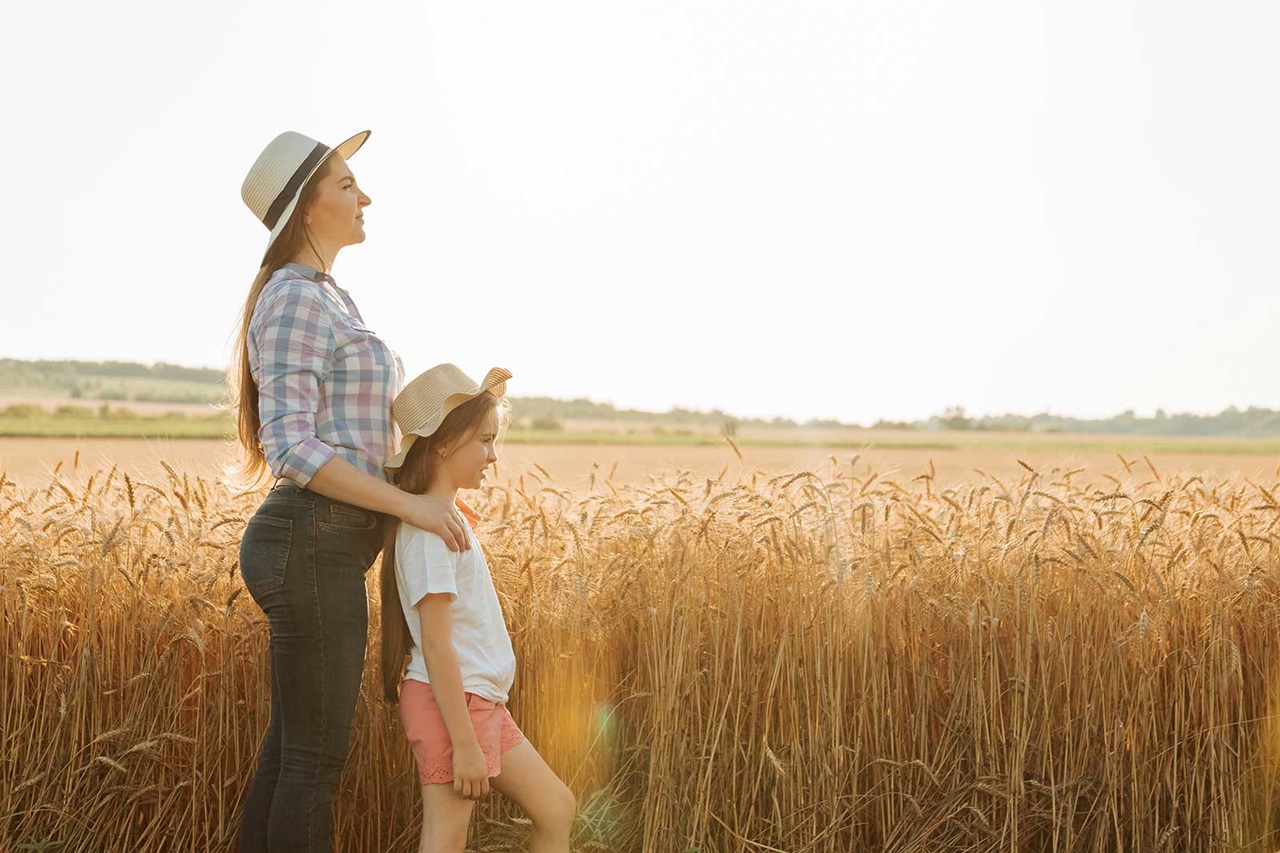 A woman and a girl wearing hats stand in a wheat field, enjoying the sunset with golden light illuminating the landscape.