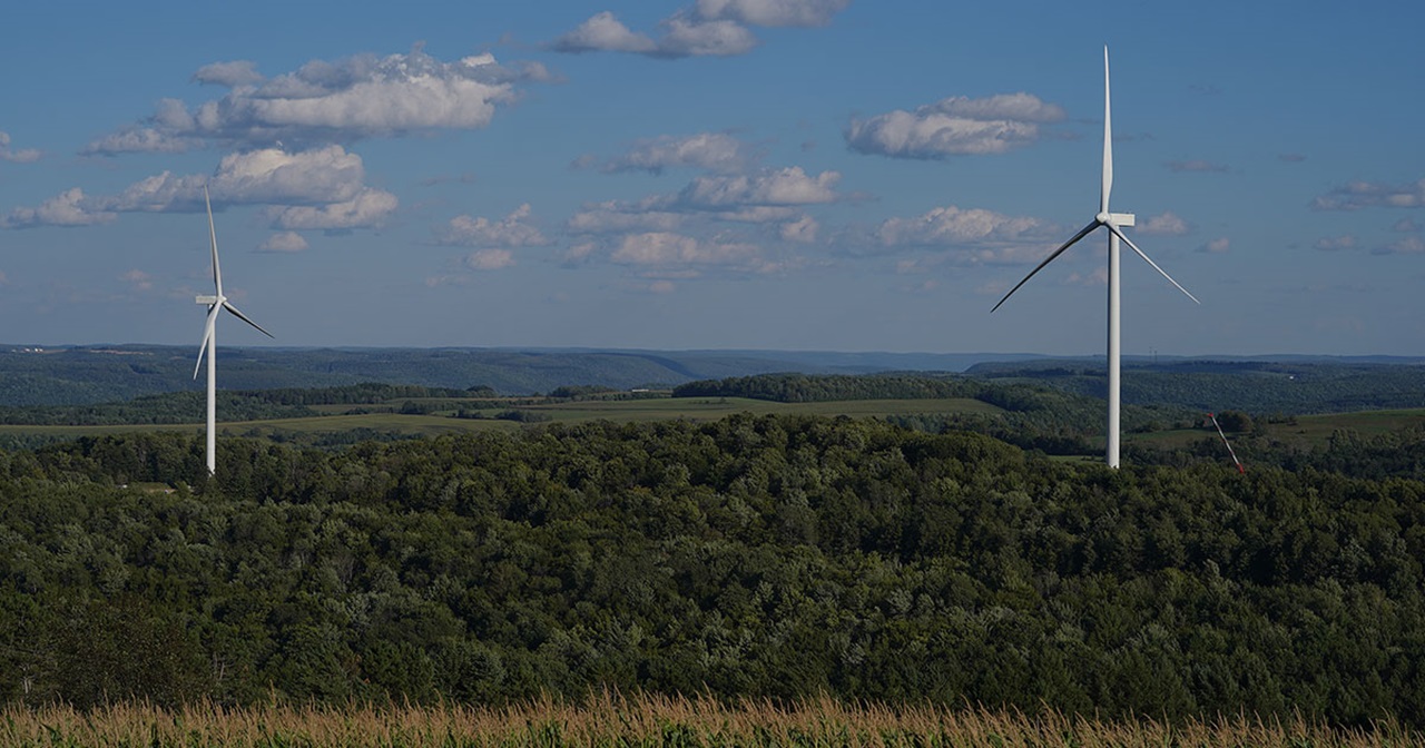 Two wind turbines stand on a green hillside under a blue sky with scattered clouds.