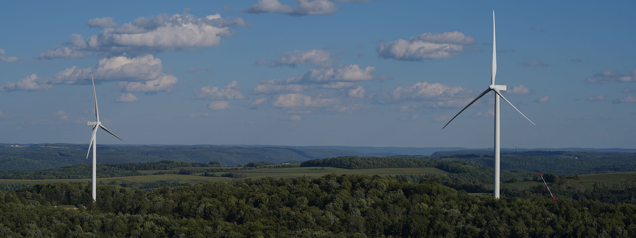 Two wind turbines stand on a lush green hillside under a clear blue sky with fluffy clouds.