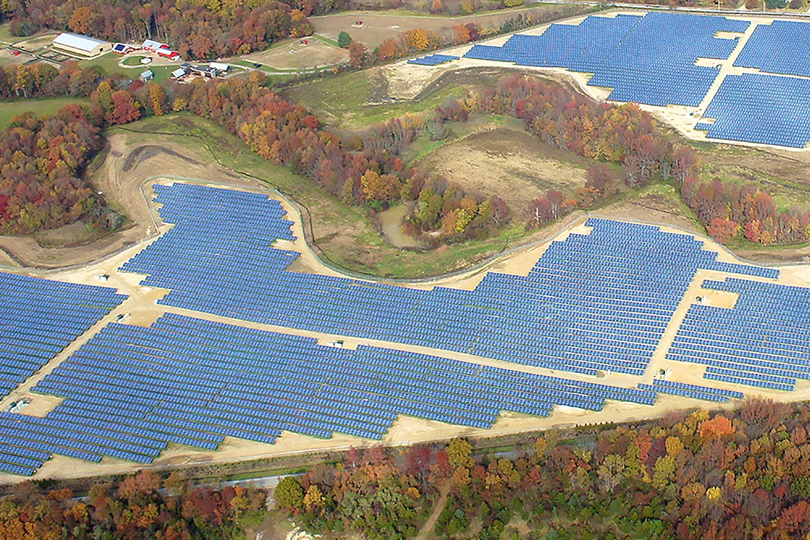 An aerial view of a large solar farm, featuring numerous blue solar panels on a landscape with colourful autumn foliage.