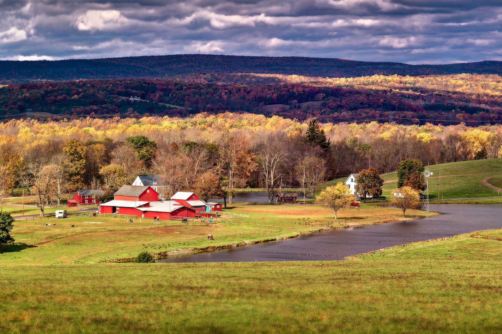 A picturesque autumn farm scene featuring red barns, a pond, and colourful trees under a cloudy sky.