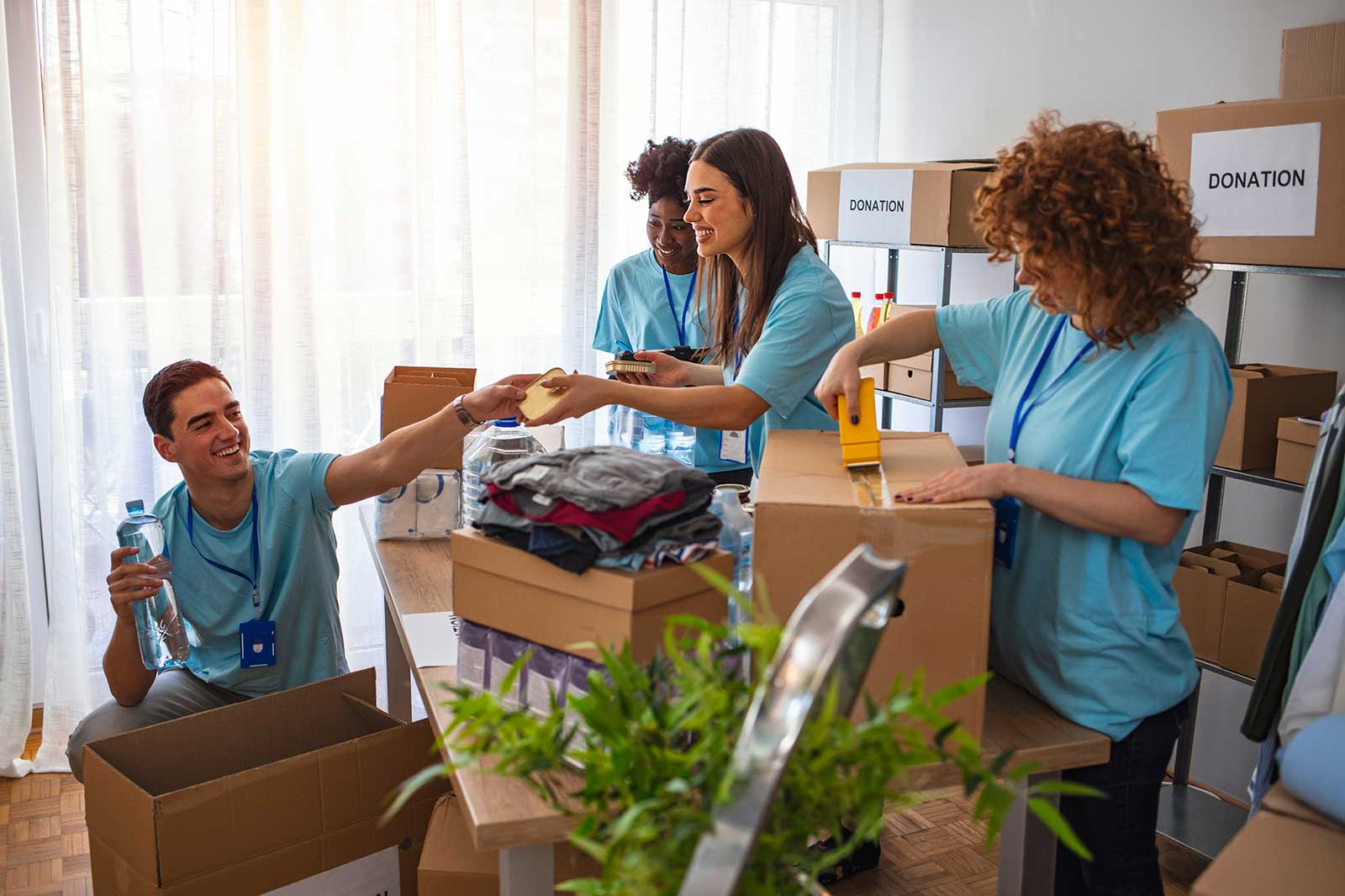 Volunteers in blue shirts are packing donations in a bright room, surrounded by boxes labelled 'DONATION' and folding clothes.