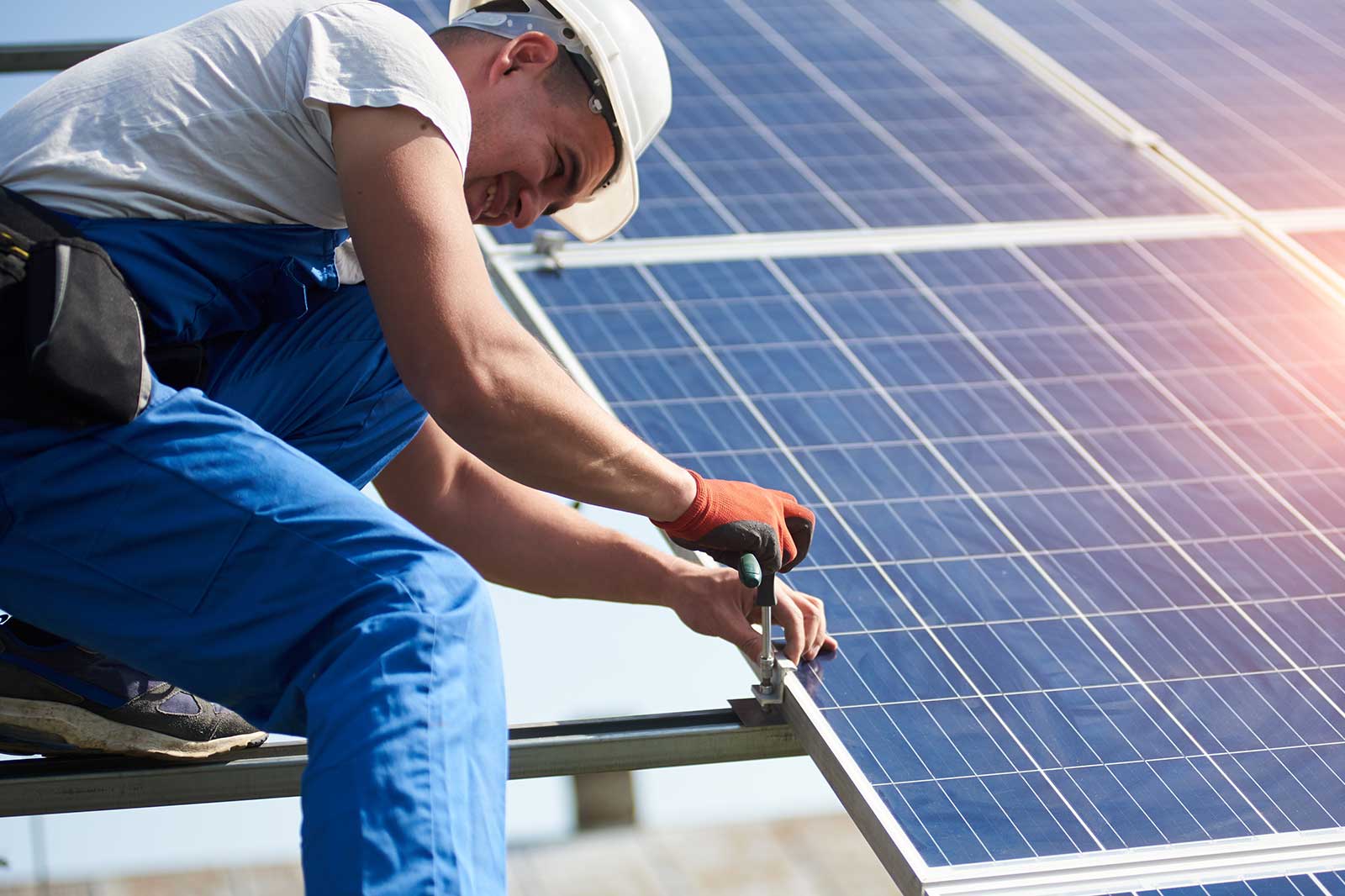 A worker in blue overalls and gloves is installing solar panels on a rooftop on a sunny day.