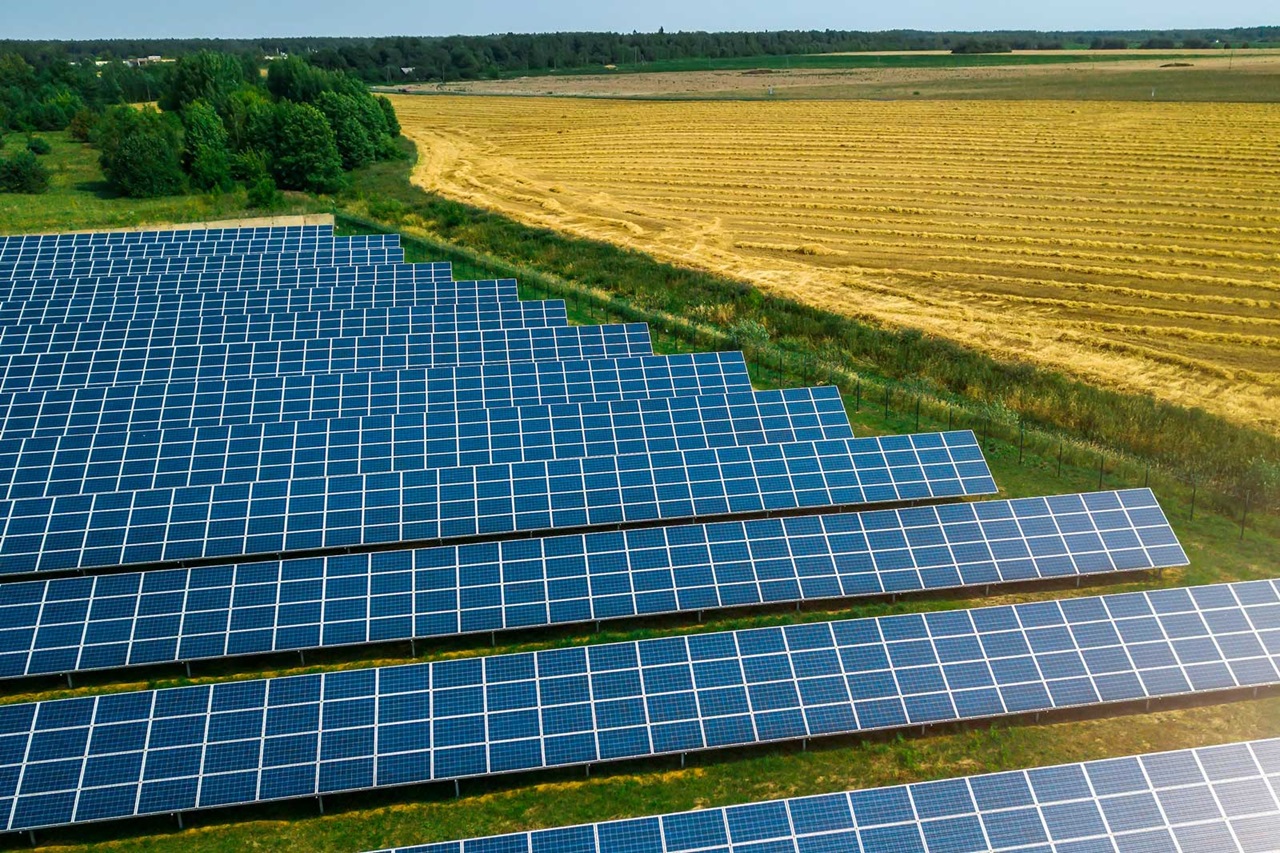 Aerial view of a solar panel farm with rows of photovoltaic panels beside a harvested field and dense green trees.