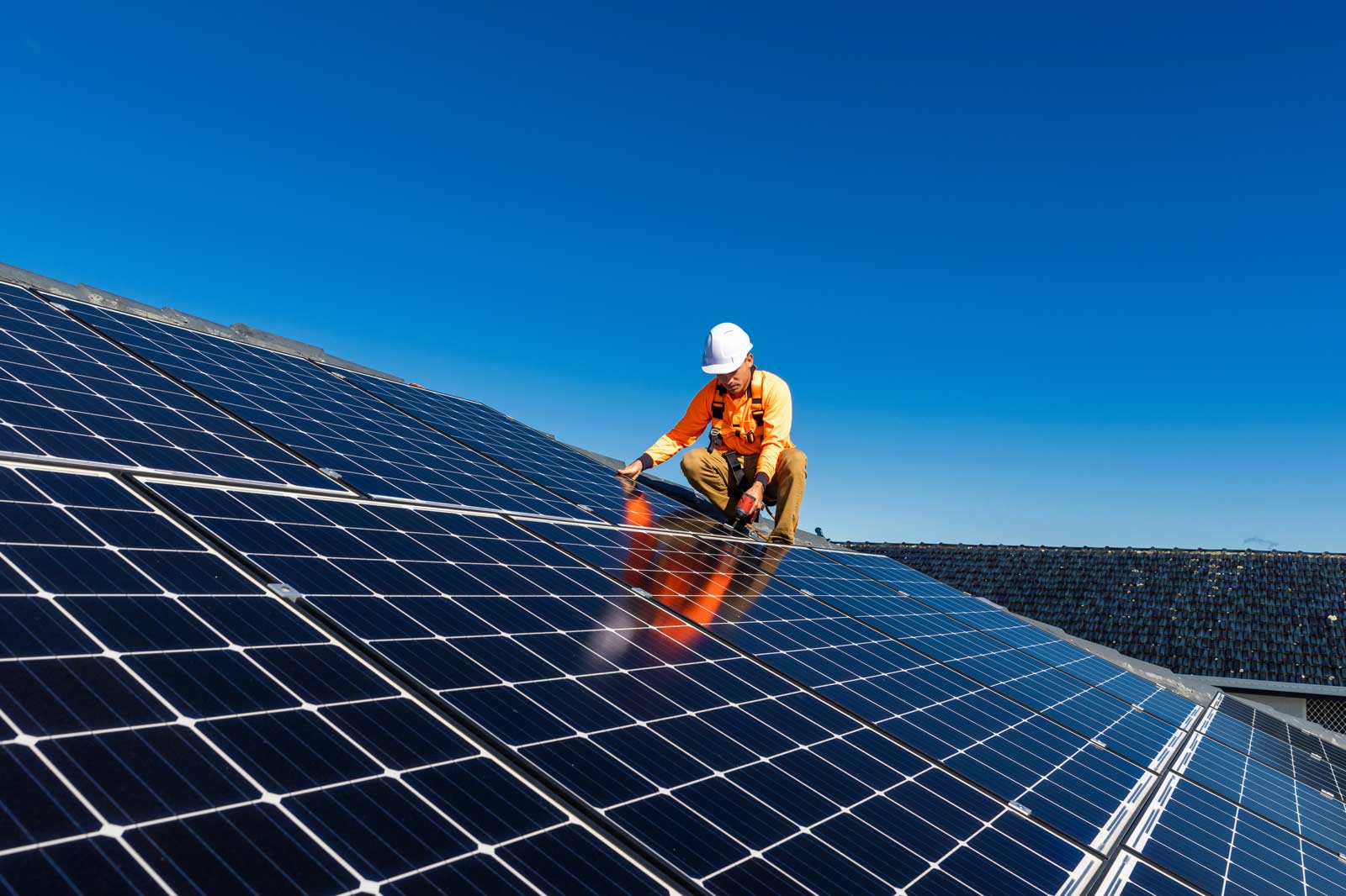 A worker in an orange safety jacket and white helmet is installing solar panels on a roof under a clear blue sky.