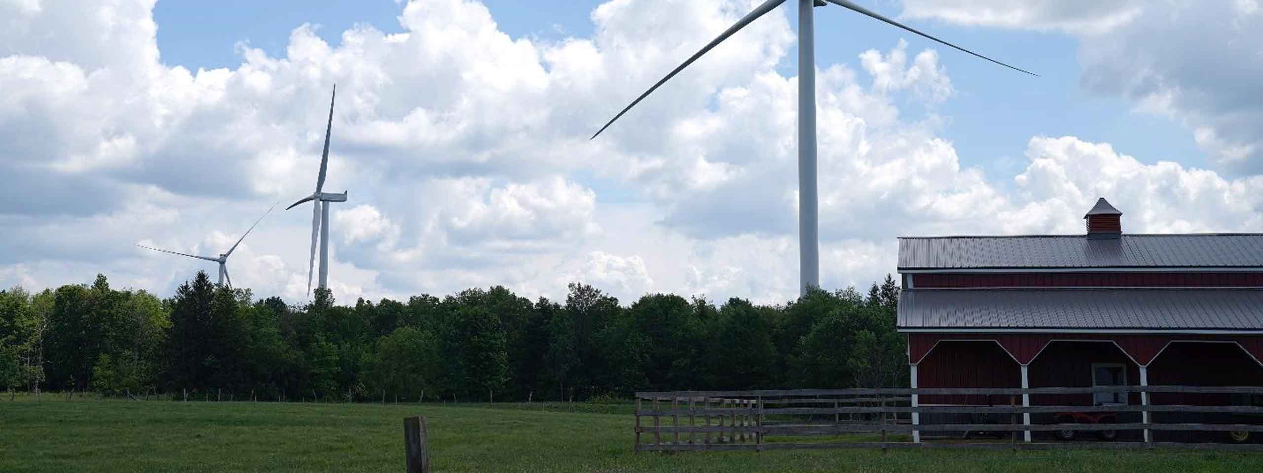 A scenic view featuring two wind turbines in a green field with a rustic red barn under a cloudy sky.