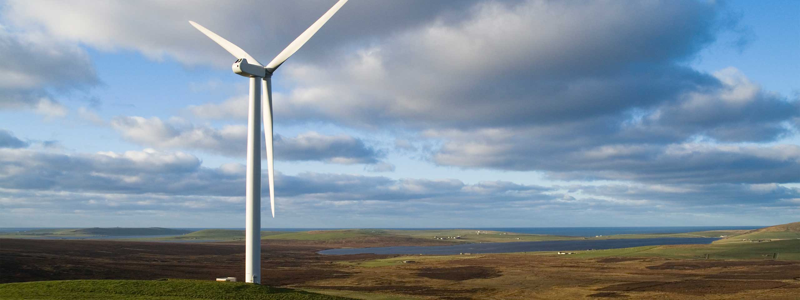 A large white wind turbine stands on green hills, surrounded by farmland and a blue sky with fluffy clouds.
