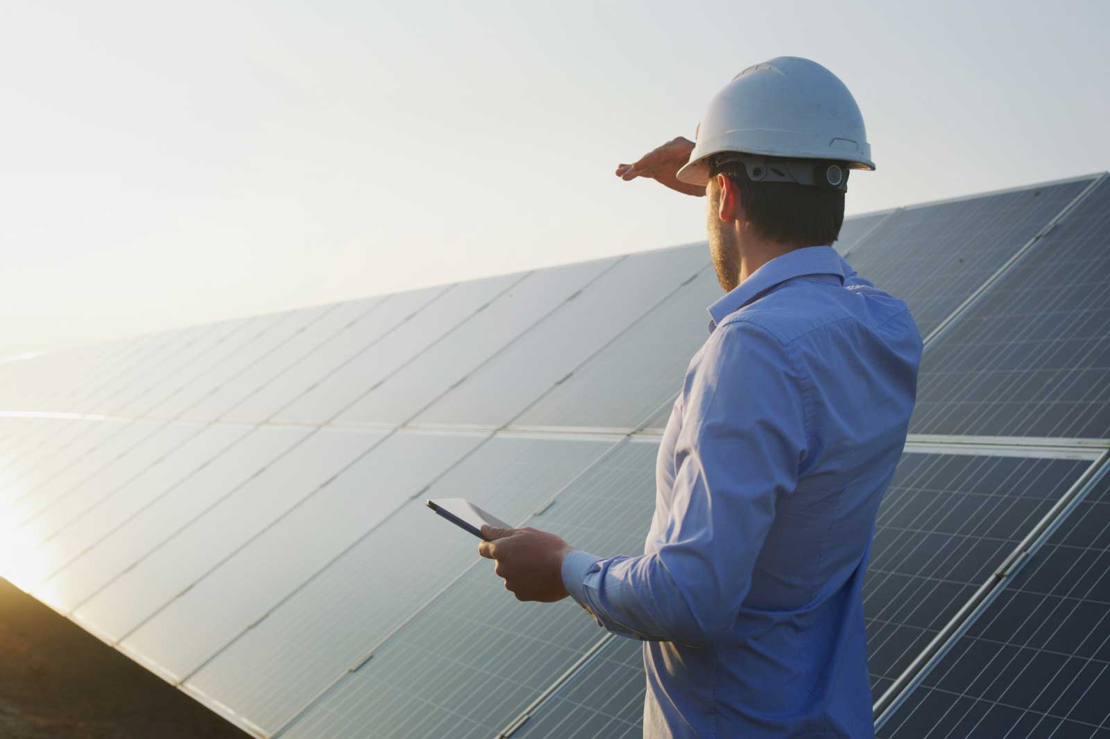 A man in a hard hat stands near solar panels, checking a device while shielding his eyes from the sun.