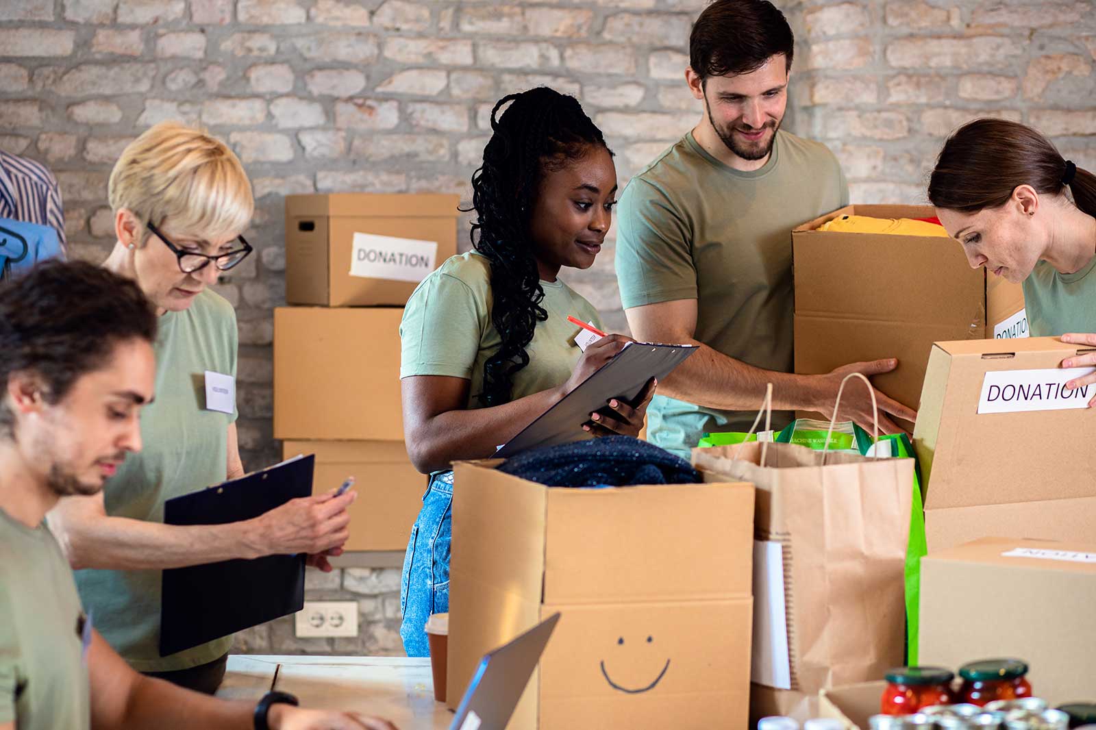 A group of volunteers organising donation boxes in a bright room with a stone wall, featuring cardboard boxes and a smiley face bag.