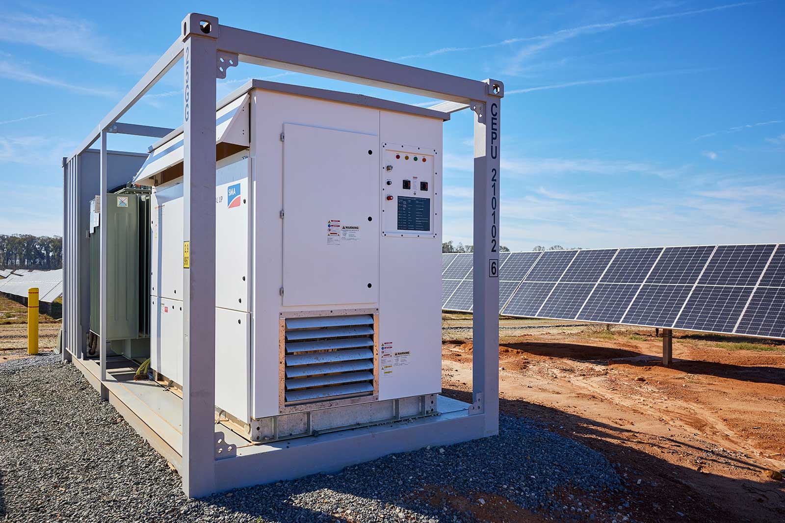 A solar inverter structure next to a field of solar panels, with clear blue skies and gravel ground.