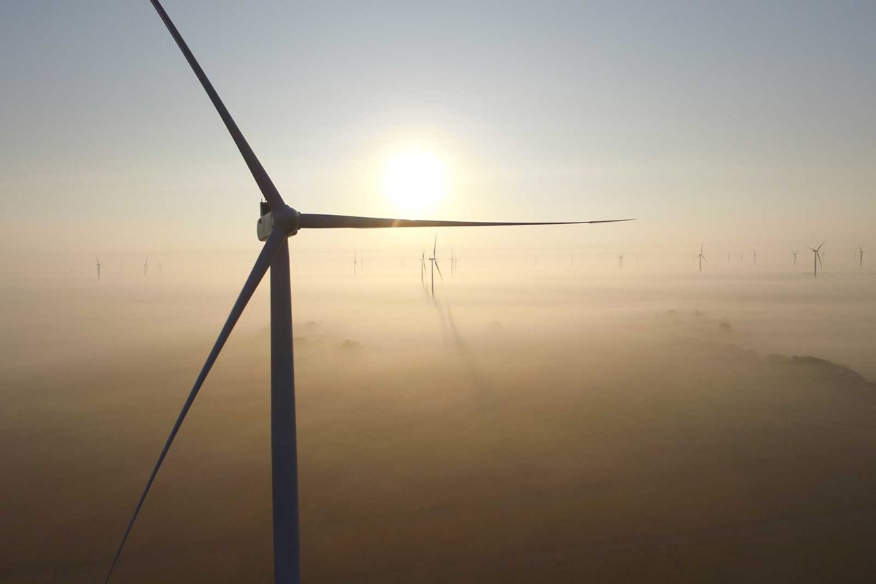A wind turbine is silhouetted against a misty landscape at sunrise, with additional turbines visible in the background.