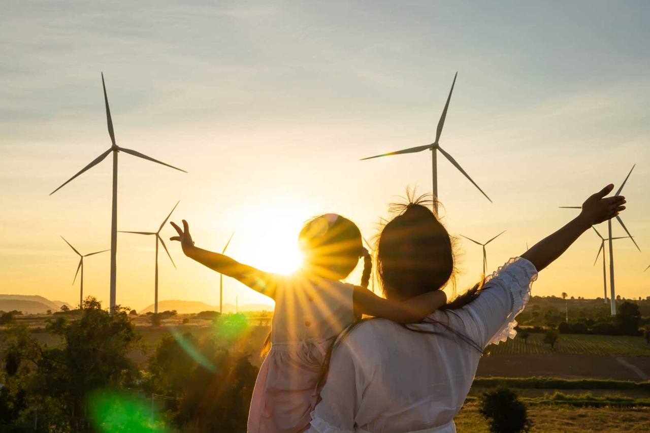 A woman and child stand together, raising their arms in joy, with wind turbines and a sunset in the background.