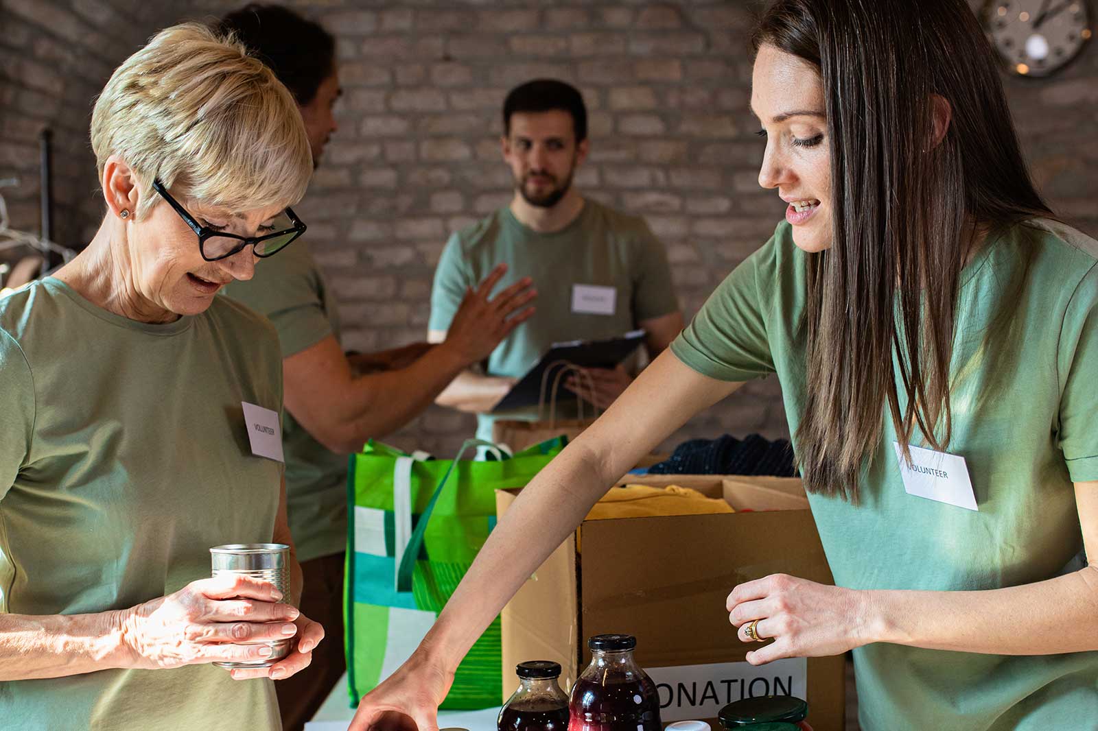 Volunteers sorting donations in a community space. They wear green shirts and handle various items, surrounded by cardboard boxes.
