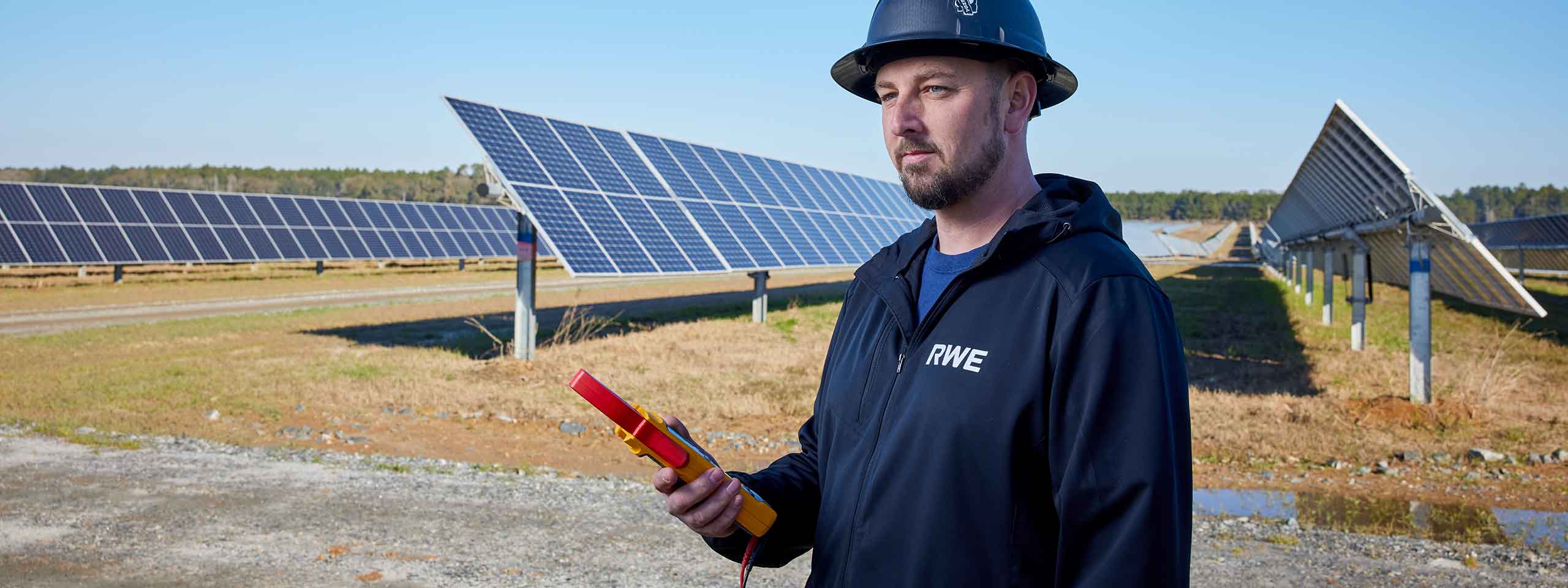 A technician wearing a hard hat and RWE jacket holds a tool in a solar panel field, with rows of solar panels in the background.