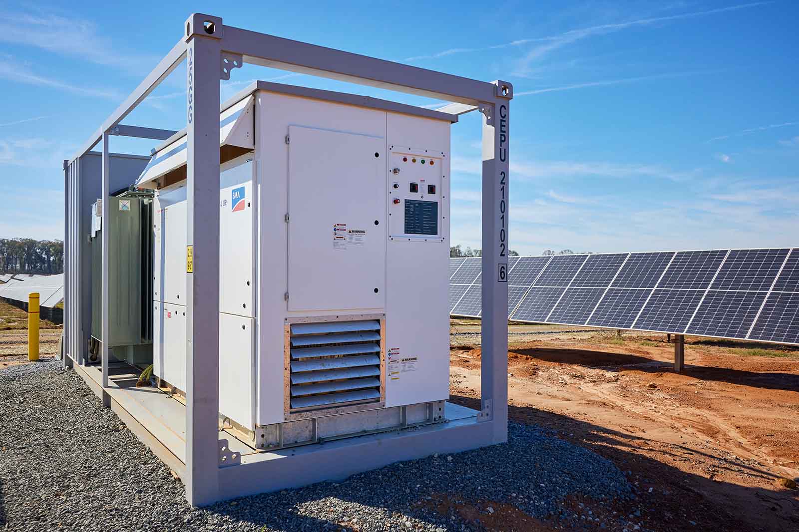 A white solar inverter housed within a metal frame, positioned next to solar panels under a clear blue sky.