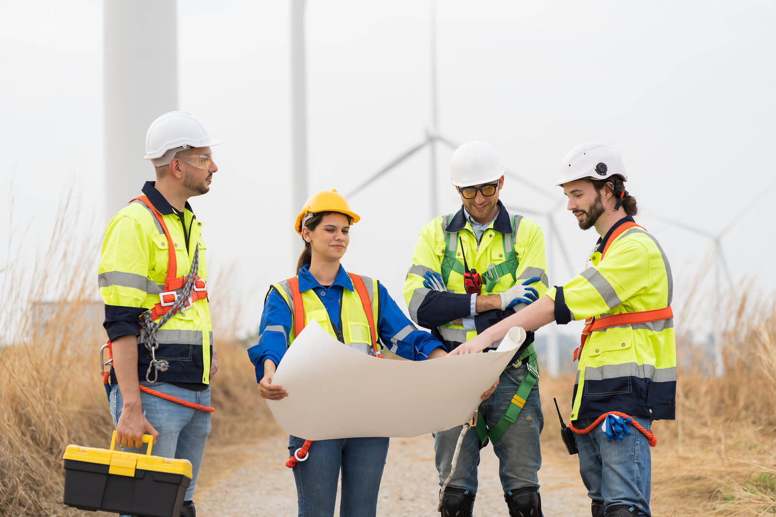 Four construction workers in safety gear consult a blueprint outdoors near wind turbines.