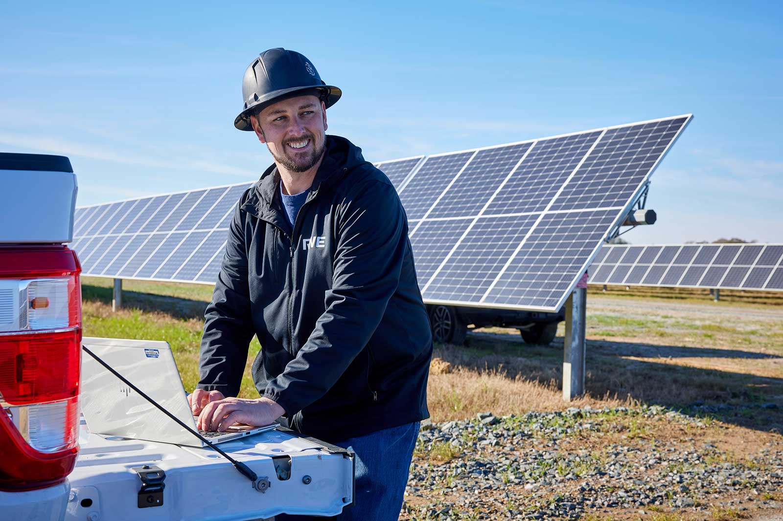 A worker in a hard hat operates a laptop on a truck's tailgate, with solar panels in the background under a clear blue sky.
