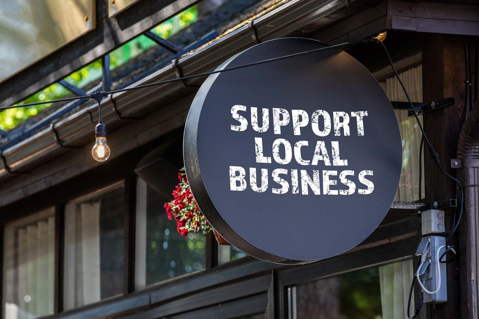A round black sign reading 'Support Local Business' hangs beside a warm light bulb near a wooden building.