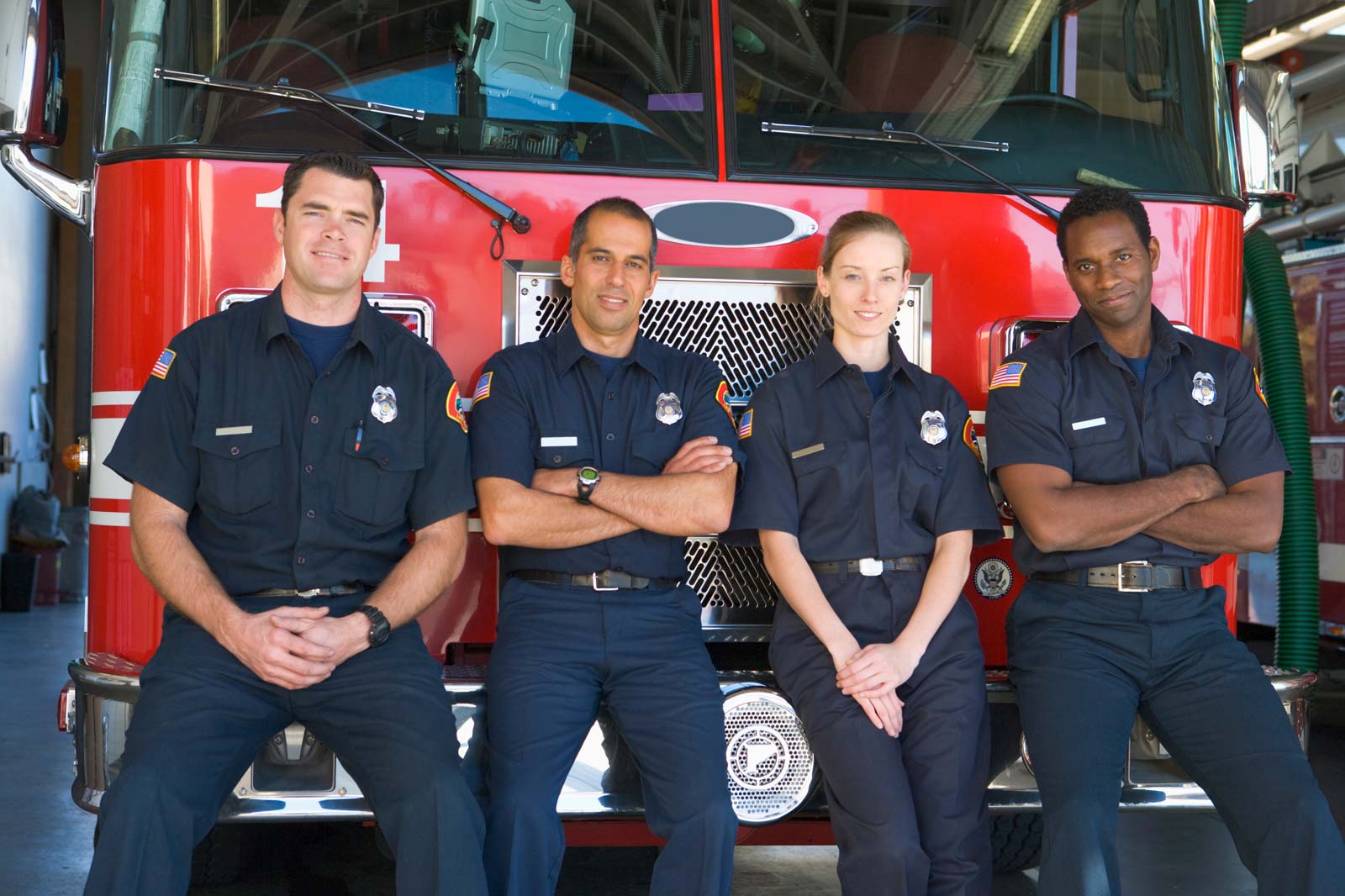Four firefighters in uniform, posing in front of a red fire truck inside a station.