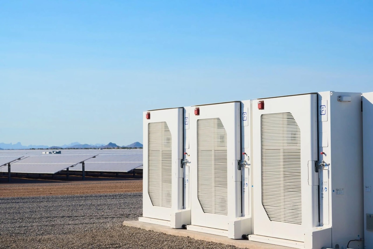 Three energy storage units positioned beside a solar panel array under a clear blue sky.
