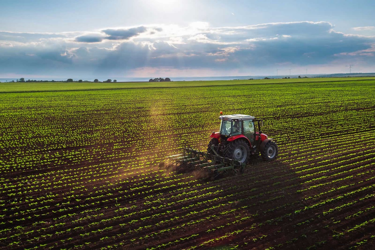 A red tractor tilling a vast green field under a cloudy sky, with sunlight streaming through.