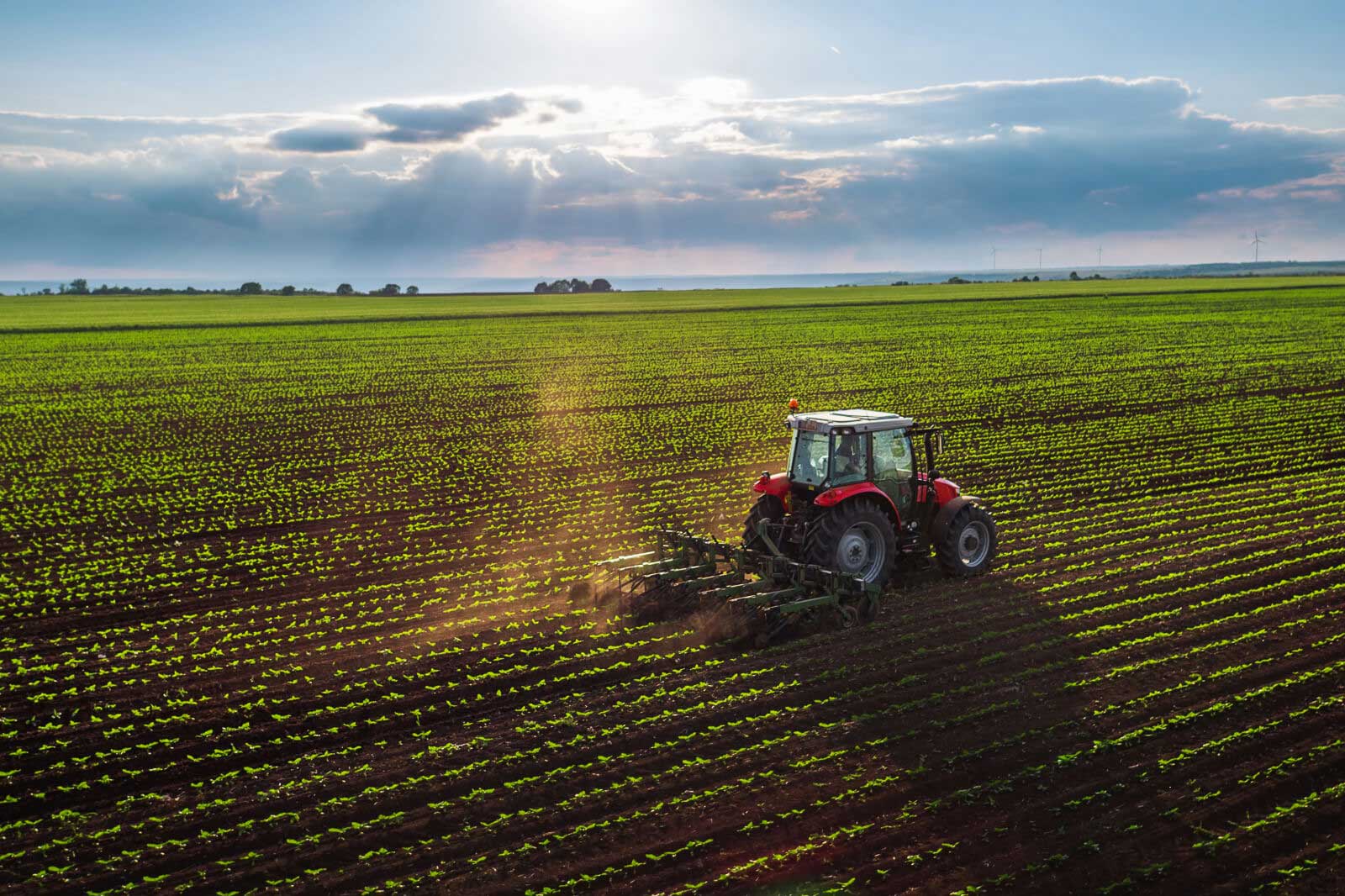 A red tractor tilling a vast green field under a cloudy sky, with sunlight streaming through.