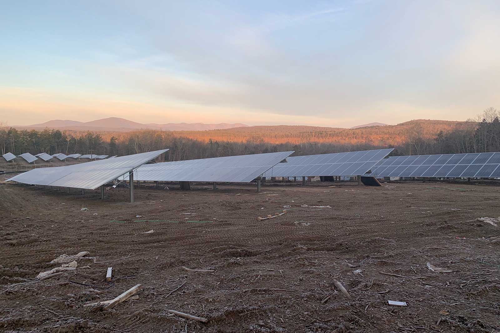 A field of solar panels set against a mountainous landscape, with a dusky sky and scattered debris on the ground.