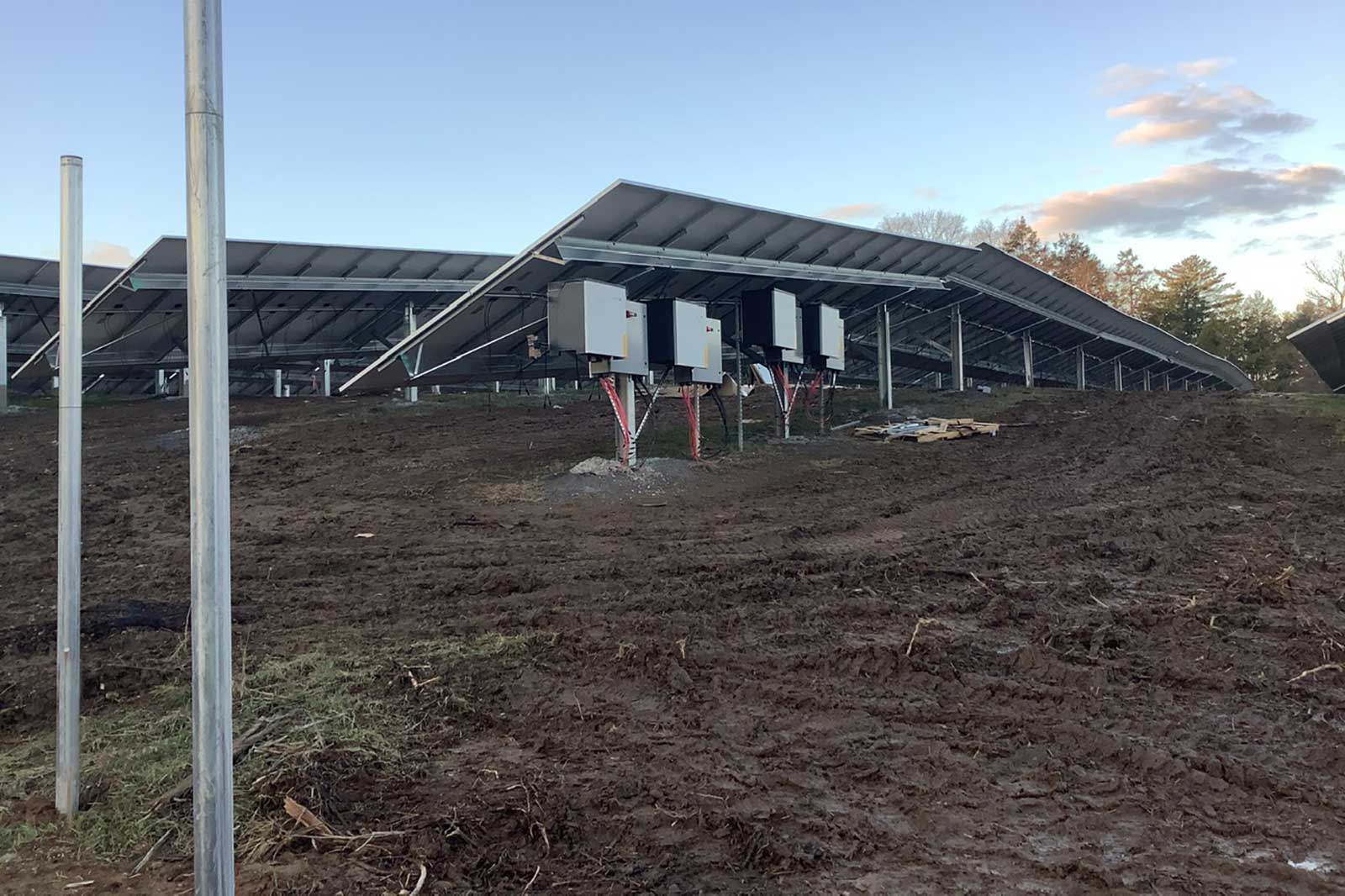 A solar panel installation with several mounted panels on metal frames and inverters on a muddy terrain under a clear sky.