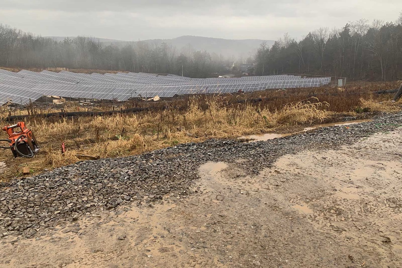 A foggy landscape with solar panel arrays, dry grass, and a gravel path. The sky is overcast, creating a muted atmosphere.