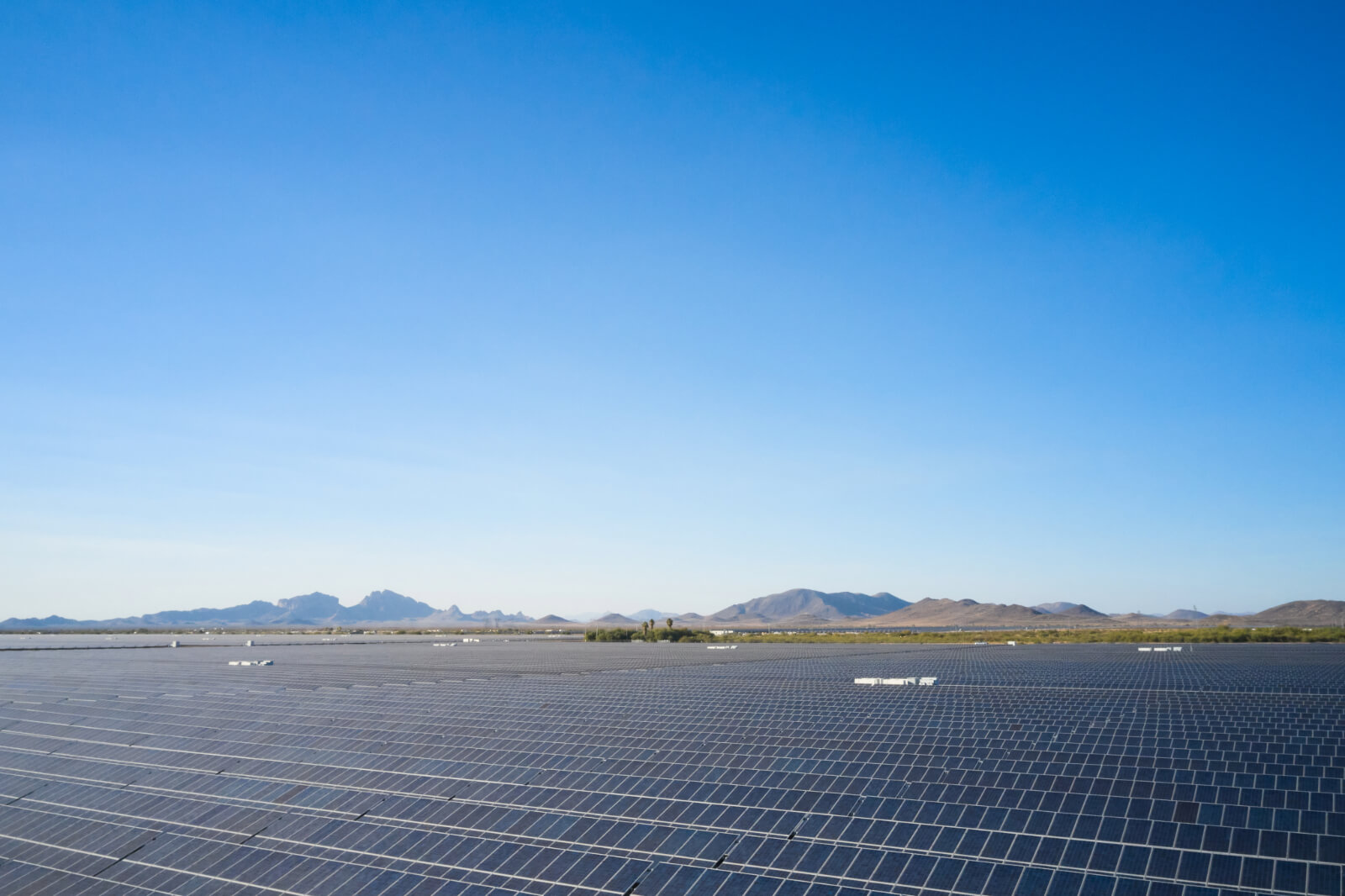 A vast solar farm with numerous solar panels installed, set against a clear blue sky and distant mountainous landscape.