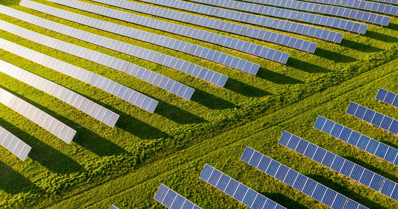 An aerial view of a solar panel field with numerous panels arranged in rows on a green grass landscape.