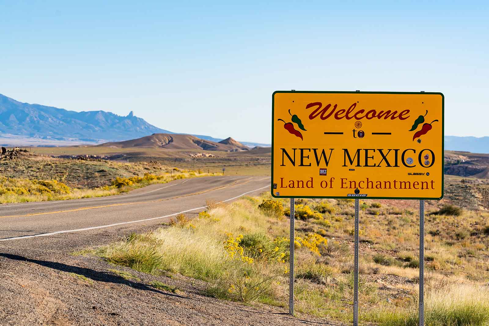 A welcome sign for New Mexico, 'Land of Enchantment', with mountains in the background under a clear blue sky.