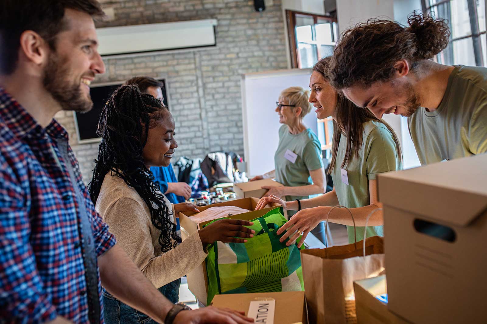 A group of people collaborating at a community event, sorting items into boxes on a table in a bright room with a brick wall.
