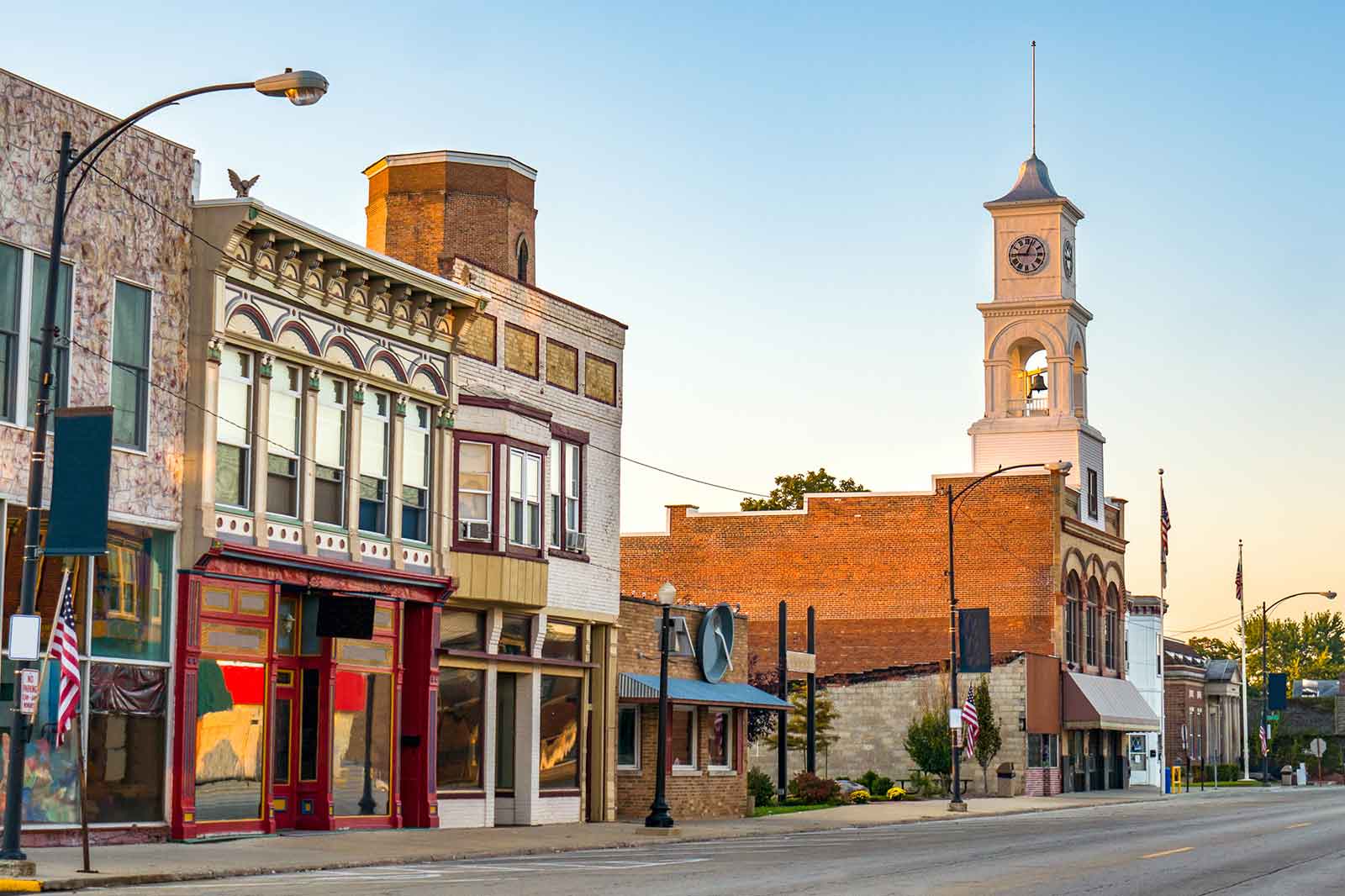 A street view featuring historic buildings, a clock tower, and American flags in a quaint town during sunrise.