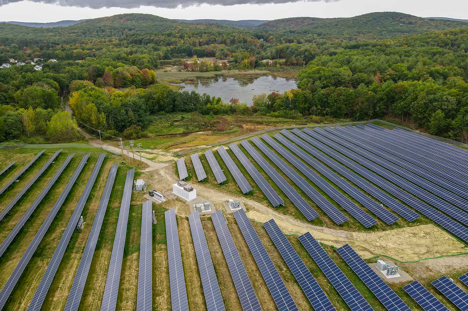 Aerial view of a solar farm with rows of solar panels, surrounded by trees and a small lake in the background.