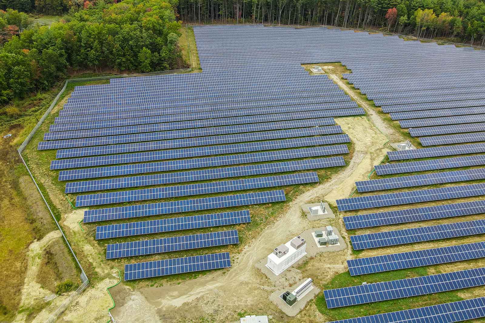Aerial view of a large solar farm with rows of solar panels, surrounded by trees, featuring a control station.