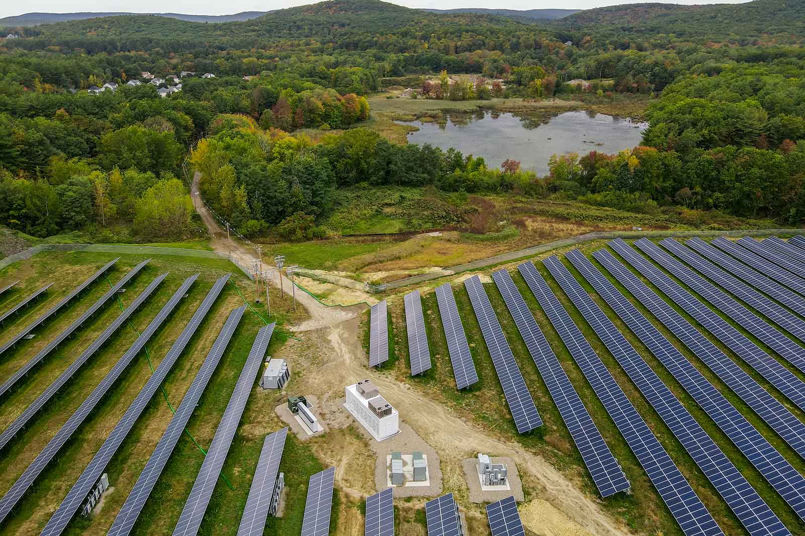 Aerial view of a solar farm with rows of solar panels, machinery, and a nearby pond surrounded by forested hills.