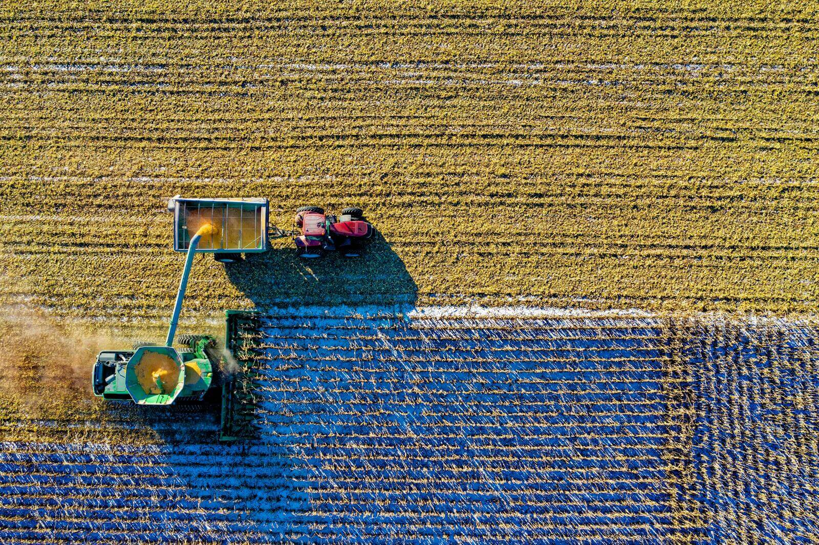An aerial view of a farm field with a combine harvester unloading into a tractor. The landscape shows harvested crops.