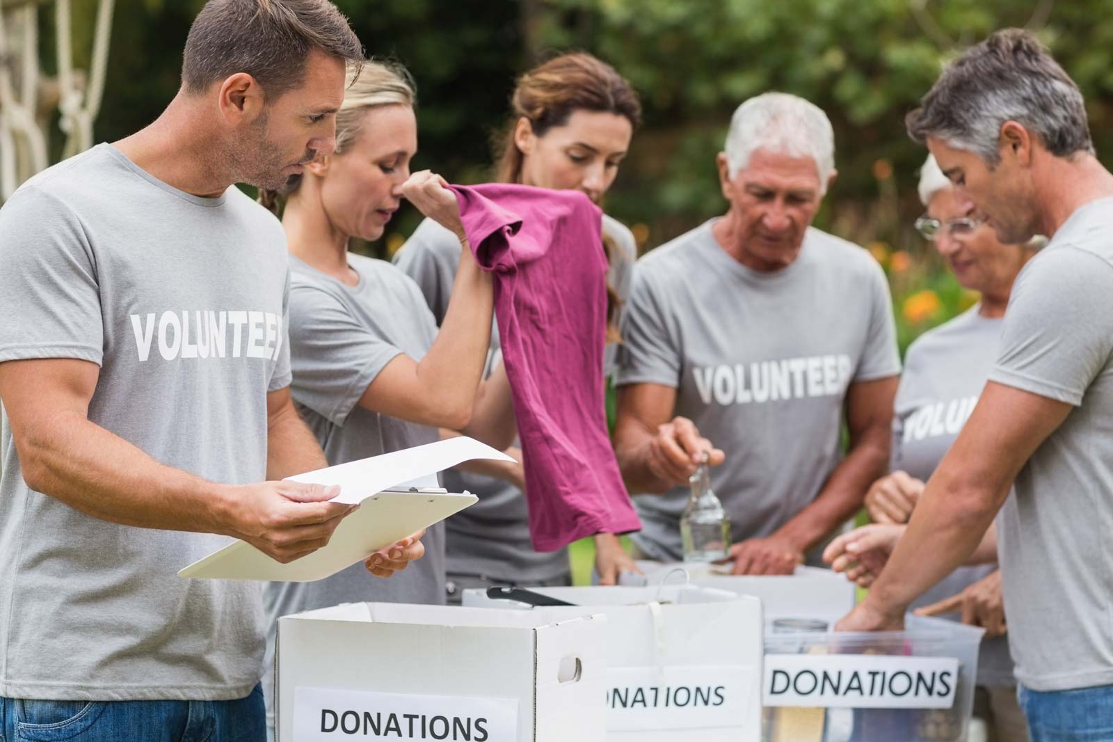 A group of volunteers in grey shirts gather around donation boxes, sorting items and discussing contributions.