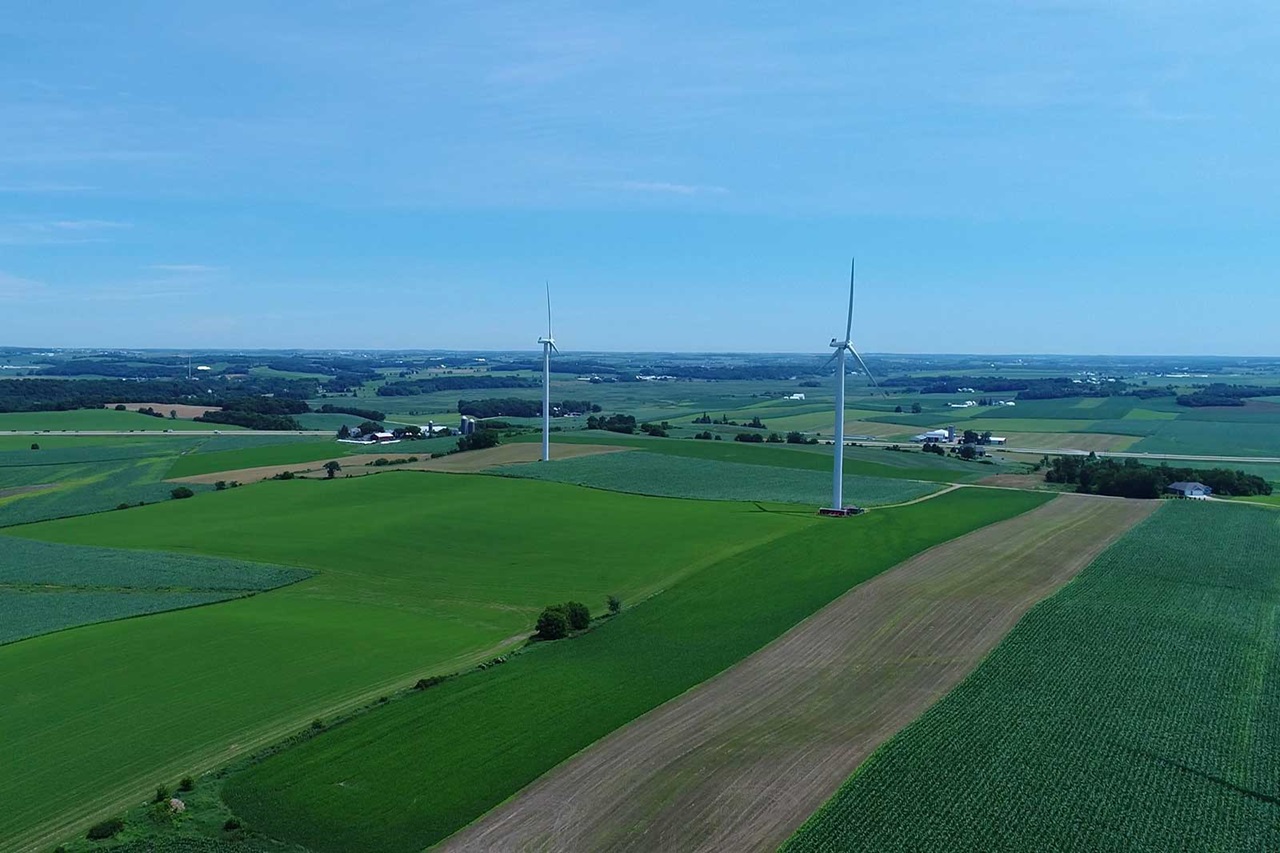 Aerial view of two wind turbines amidst lush green farmland under a clear blue sky.